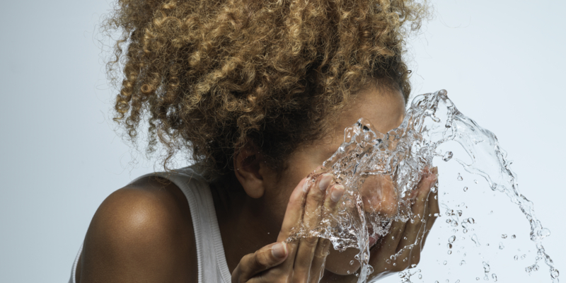 Young woman with dark skin and curly hair splashing face with fresh water against blue background
