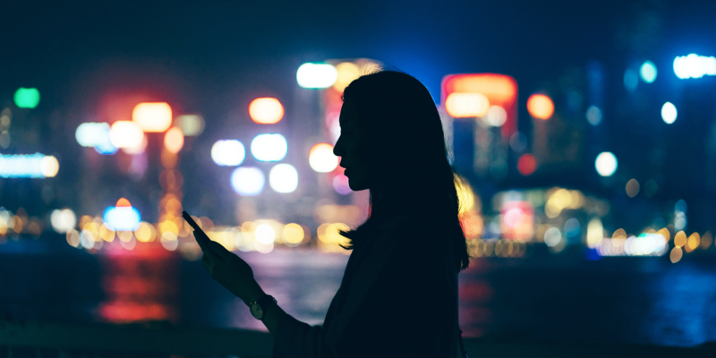 Silhouette of woman using mobile phone while standing against illuminated and multi-coloured cityscape at night